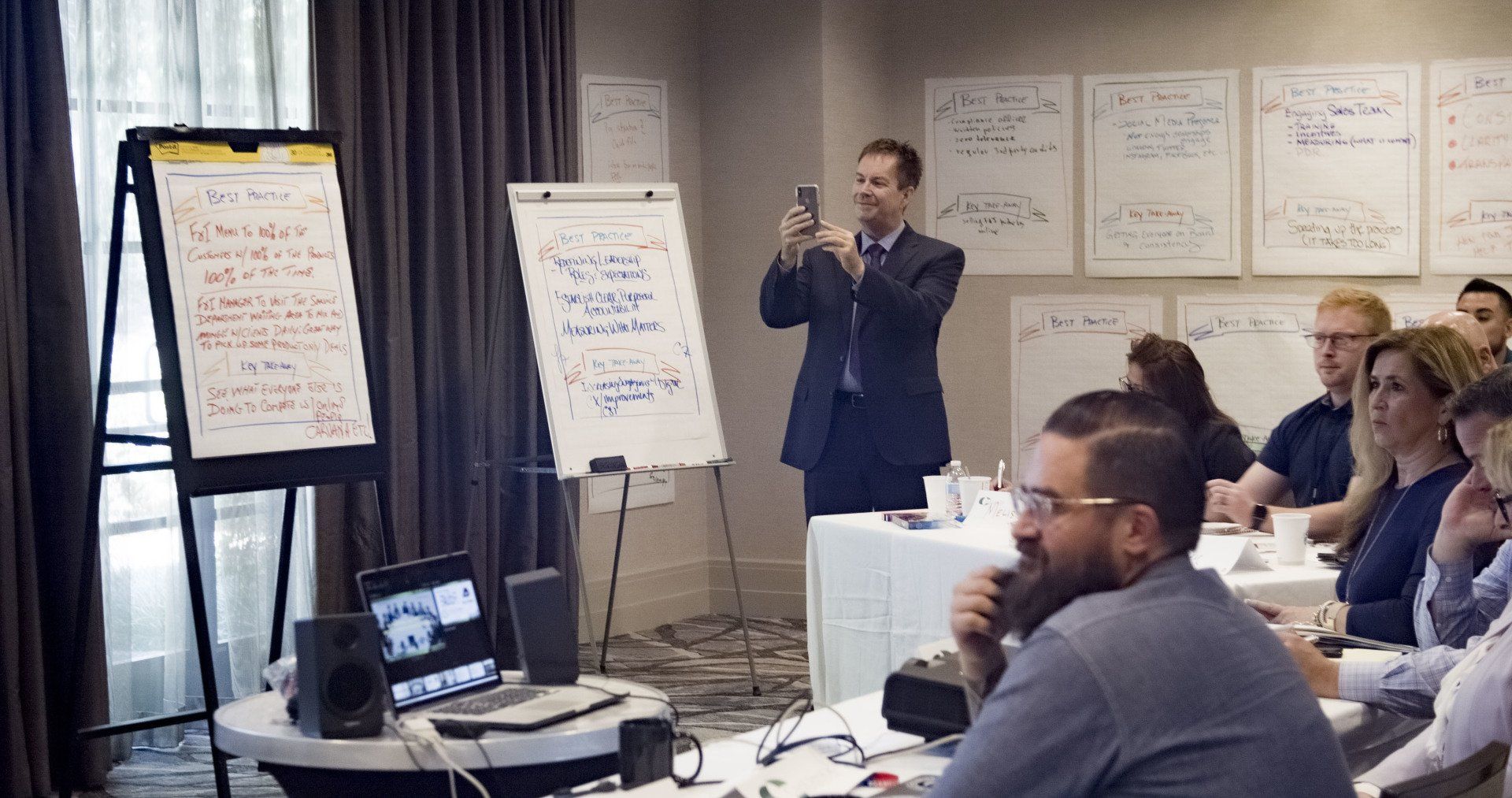Man taking photo of notes on a whiteboard during a presentation; audience seated, laptop and speakers visible.