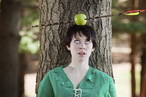 Woman with apple on head pierced by arrow in front of tree, eyes rolled up, wearing green shirt.