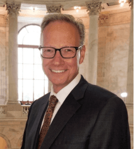 Man in suit and glasses smiles in ornate building.