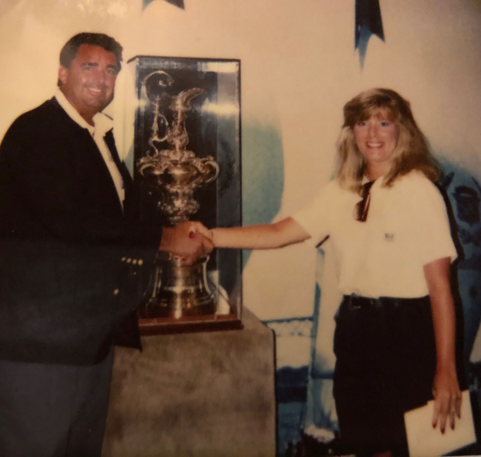 Man in suit shakes hands with woman in front of a trophy, possibly a sailing cup.