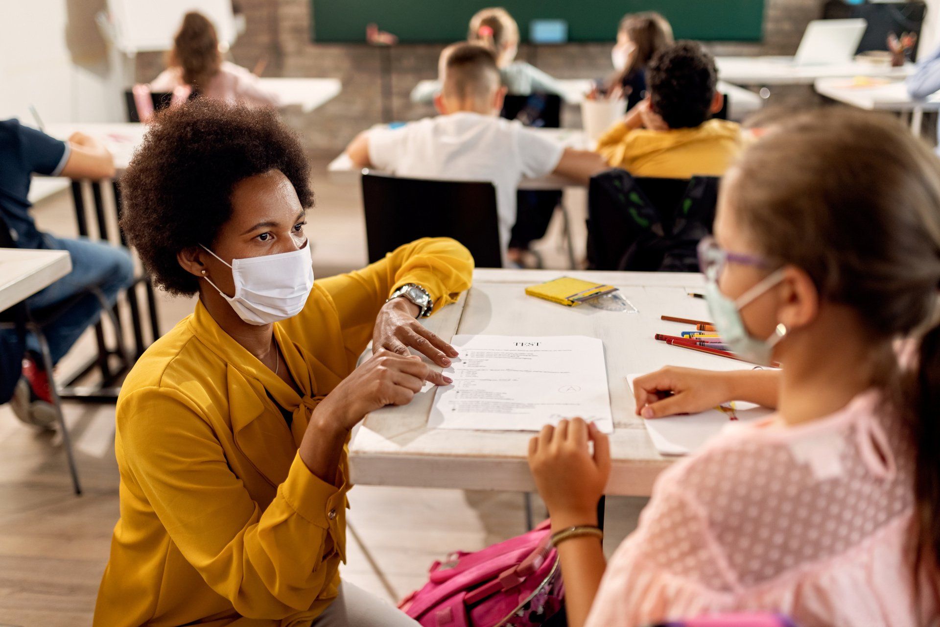 A teacher wearing a mask is talking to a student in a classroom.