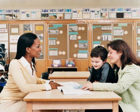 A teacher sits at a desk with two students in a classroom