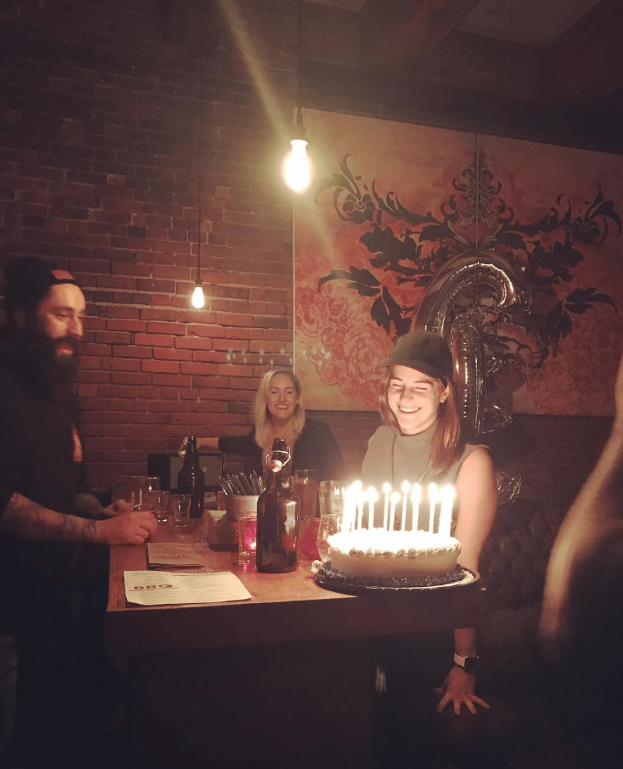 Woman holding birthday cake with lit candles; smiles; friends in a dimly lit bar with exposed brick wall and a mural.
