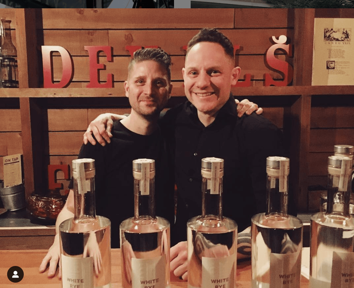 Two men, smiling, pose behind a bar with bottles. Wooden shelf backdrop with 