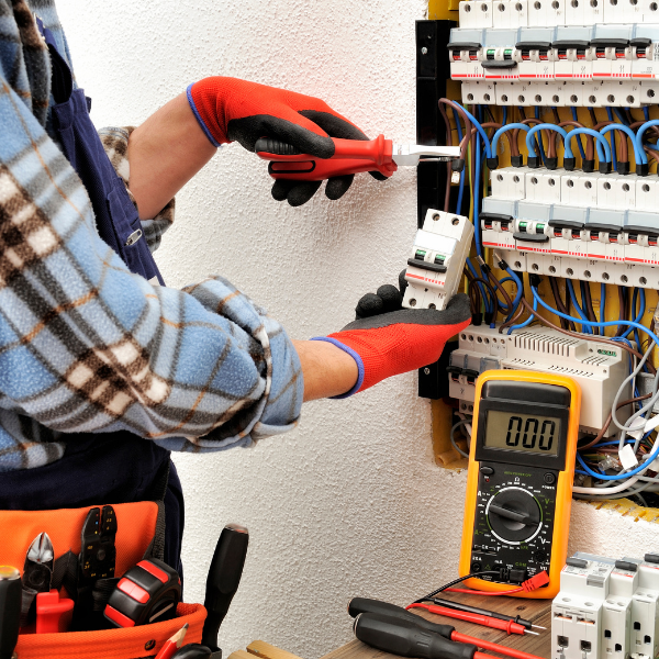 Electrician testing a circuit breaker panel with a multimeter and screwdriver, wearing red gloves.