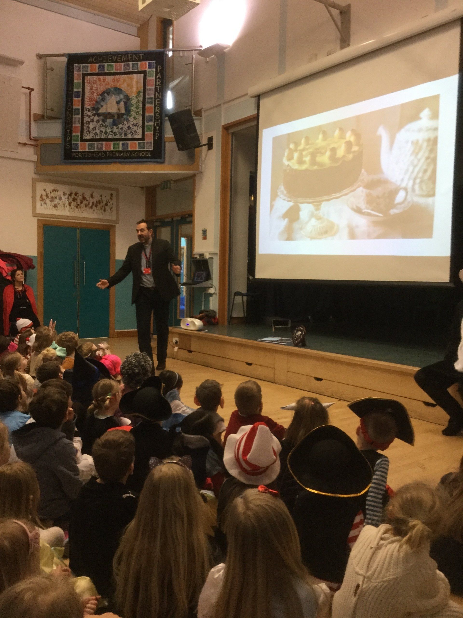 Man lecturing children, projecting image on a screen, indoors. Children sit on the floor, some in costume.