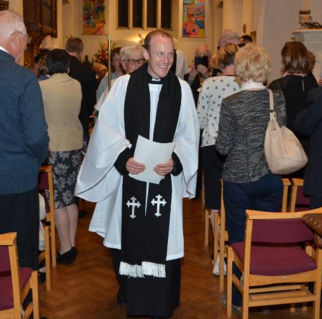 A clergyman smiling, walking down an aisle in a church, holding papers; people around.