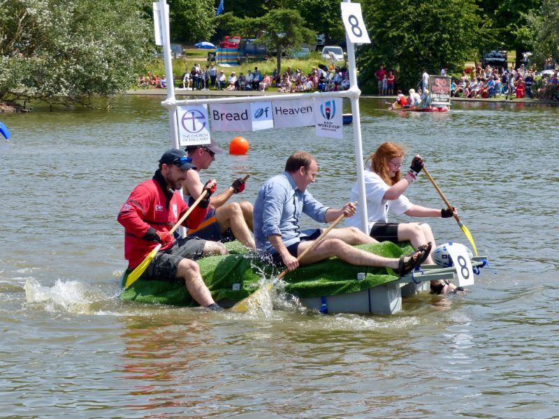 Four people paddling a homemade raft in a river, with a banner and numbered sign.