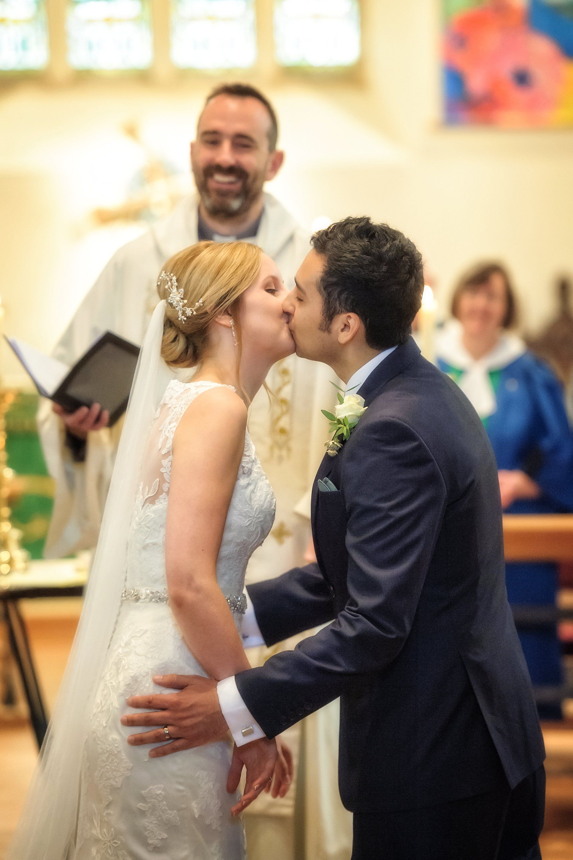 Bride and groom kiss at altar during wedding ceremony, officiant smiles in background.