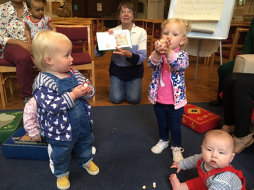 Toddler group in a church. A woman reads a book to babies, some play with blocks.