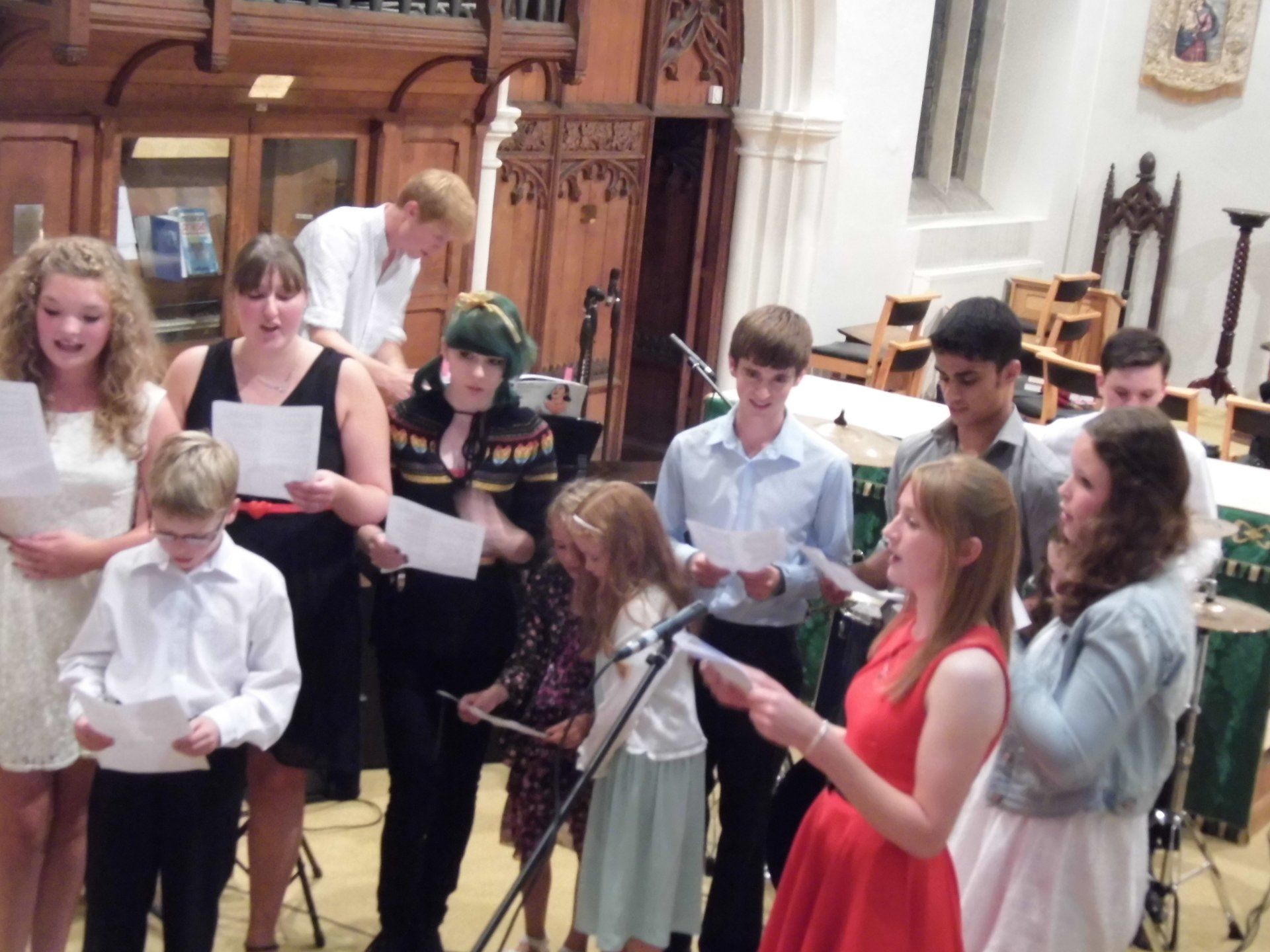 Group of people singing, holding sheet music, in a church. Microphones and instruments present.