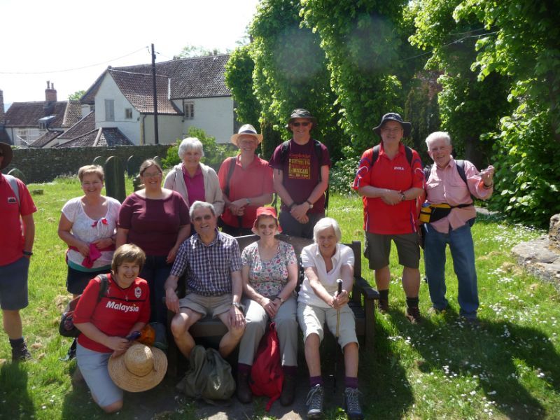 Group of people smiling outdoors near a building with a lush green backdrop. Some are seated on a bench.