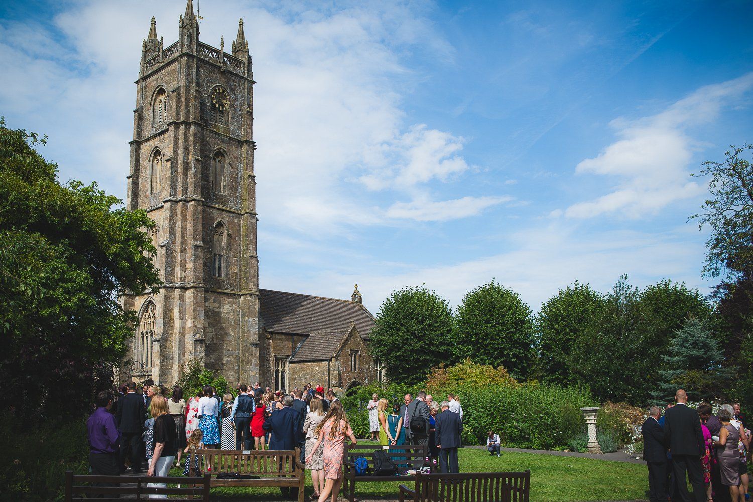 Wedding guests gather on a lawn in front of a stone church with a tall tower under a blue sky.