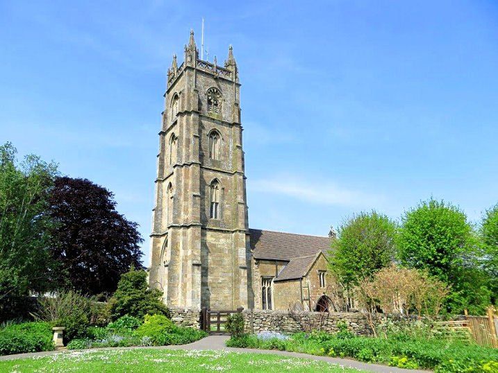 Stone church with tall tower, under a blue sky, surrounded by green trees and a garden.
