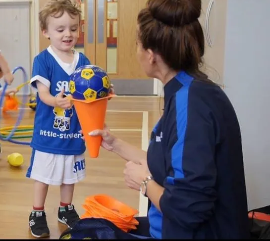 Young boy in blue jersey hands a soccer ball on a cone to a woman in a blue tracksuit indoors.