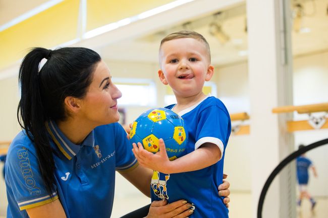 Woman and young boy in blue shirts, holding a soccer ball, smiling in a gym.