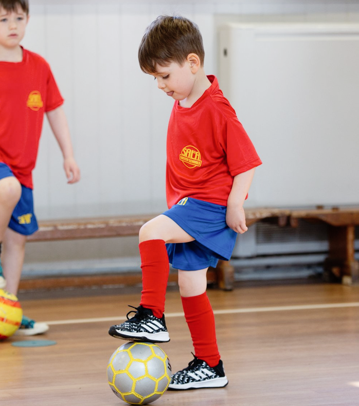 Boy in red shirt and blue shorts, foot on soccer ball. Indoors, other child nearby.