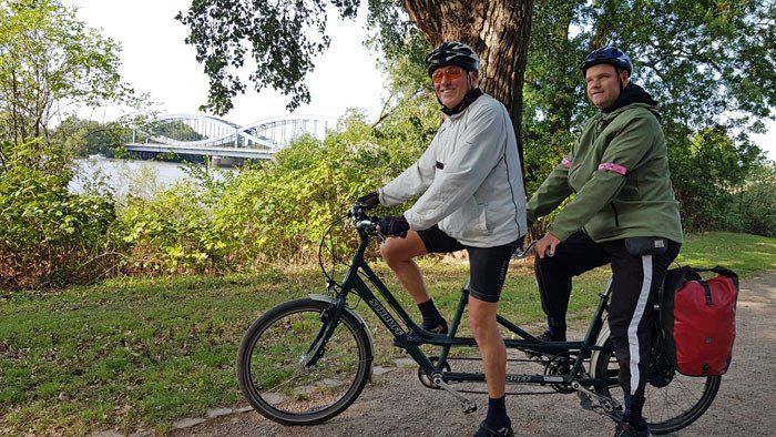 Das Foto zeigt Arno und Rudi auf dem Tandem im Park Entenwerder an der Elbe. Zwischen den Sträucher im Hintergrund sieht man die Bögen der Elbbrücke der Autobahn