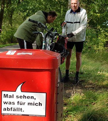 Foto von Rudi und Arno. Davor ein großer, roter Mülleimer mit dem Spruch