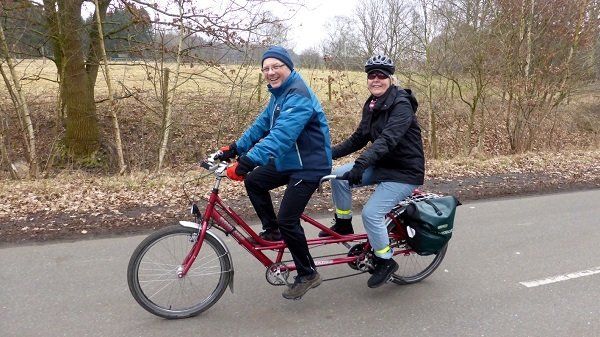 Barbara Hömbeg und ihr Pilot Ulrich Kudlek fahren auf einem Tandem eine Straße entlang, die von einer Wiese und Bäumen gesäumt ist