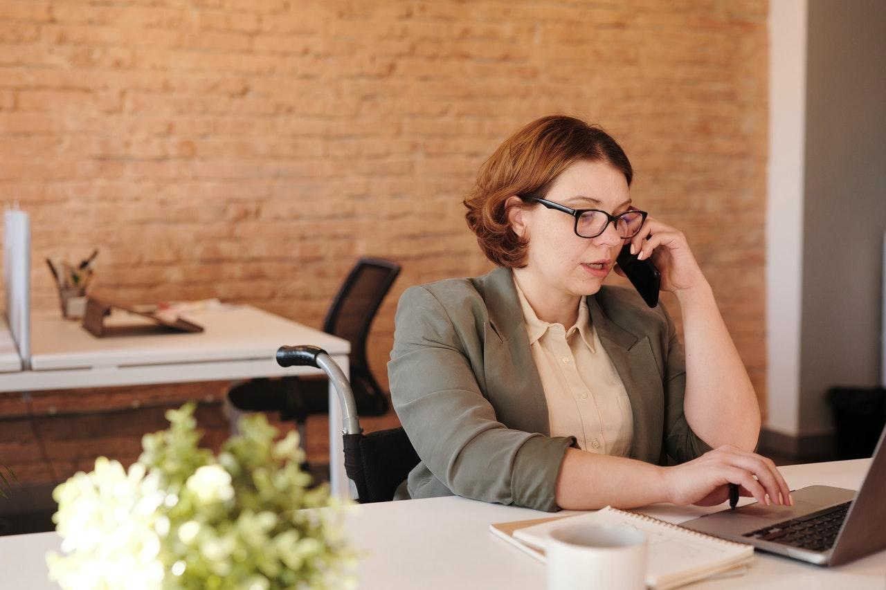 A woman in a wheelchair is sitting at a desk talking on a cell phone.