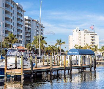 Boats docked at marina with condos and palm trees under a blue sky.