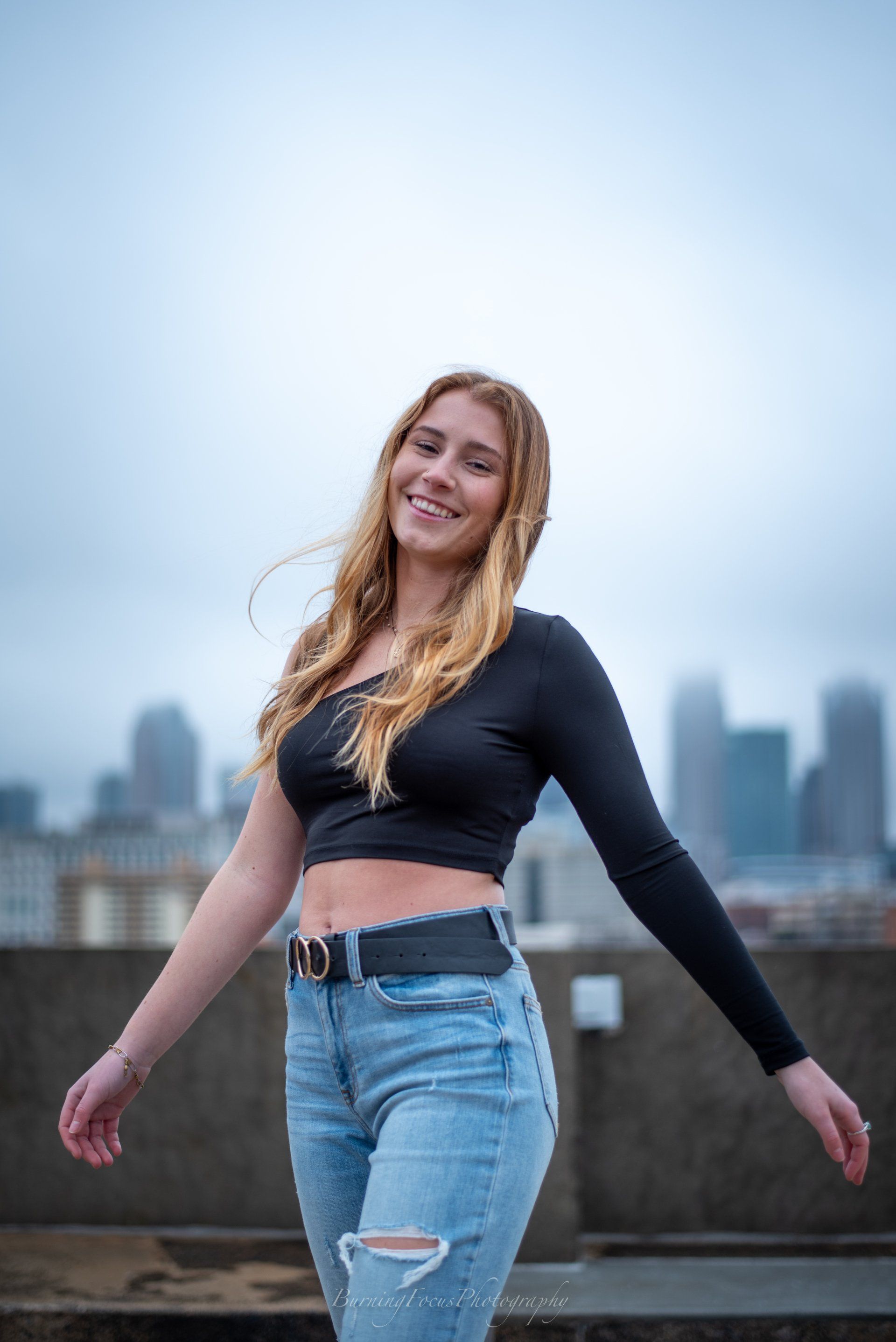 A woman in a black crop top and blue jeans is standing on a rooftop with her arms outstretched.