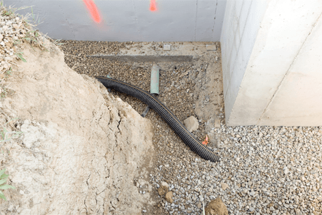 Excavation with drainage pipe next to a concrete wall and earth bank.
