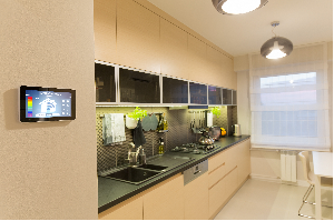 Kitchen interior with smart home control tablet on the wall, overlooking counter, sink, stove, cabinets, and window.