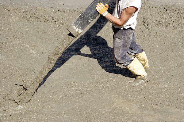 Construction worker pouring wet concrete from a black chute, outdoors on a sunny day.