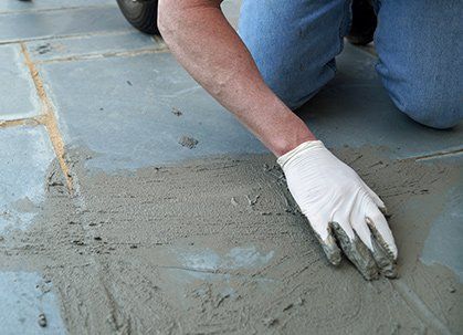 Gloved hand smoothing grout between gray stone patio tiles.