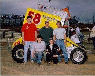 A racing team poses with a yellow sprint car numbered 58. Men stand and kneel in front of the car, outside at the track.