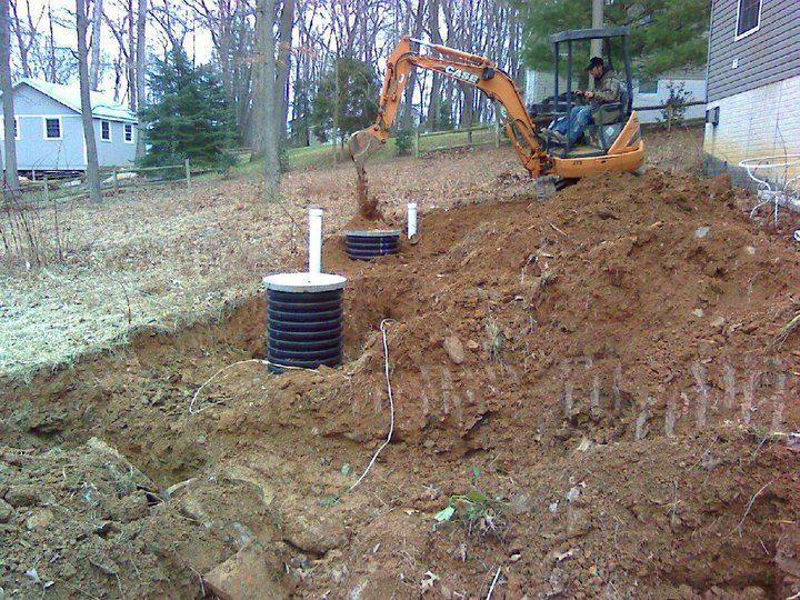An excavator digs near two black septic tanks, house in the background. Operator in the machine. Brown soil.