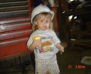 Child in a white hard hat and shirt, holding a tool, standing next to a toolbox.