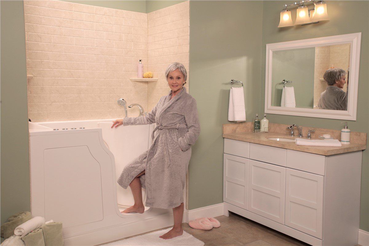 Woman in a gray robe steps into a walk-in tub in a bright bathroom. She has a towel on her head.