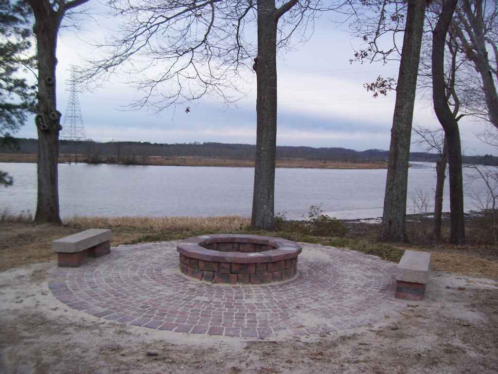 Fire pit with brick circle, two benches, overlooking a lake, trees surrounding.