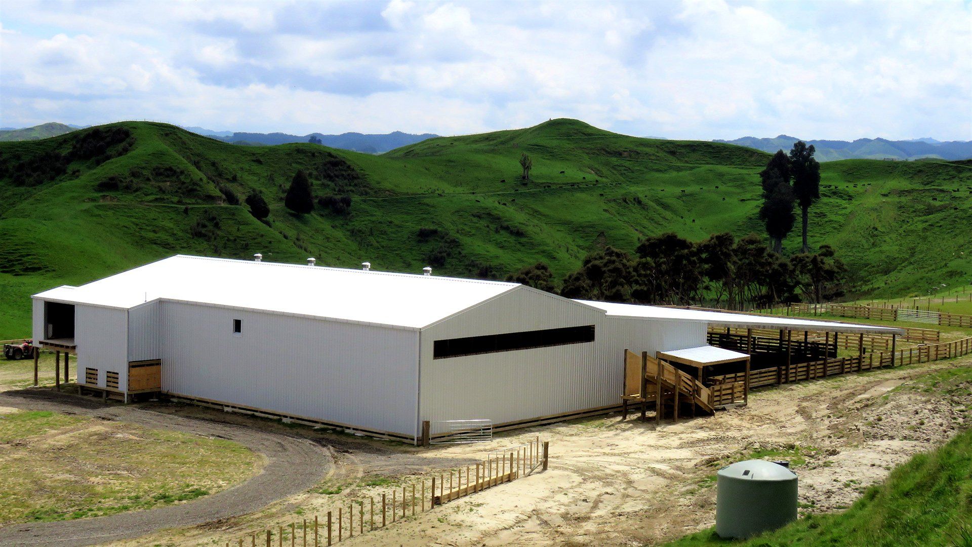 A large white agricultural building sits in a hilly green landscape with a fenced livestock area nearby.