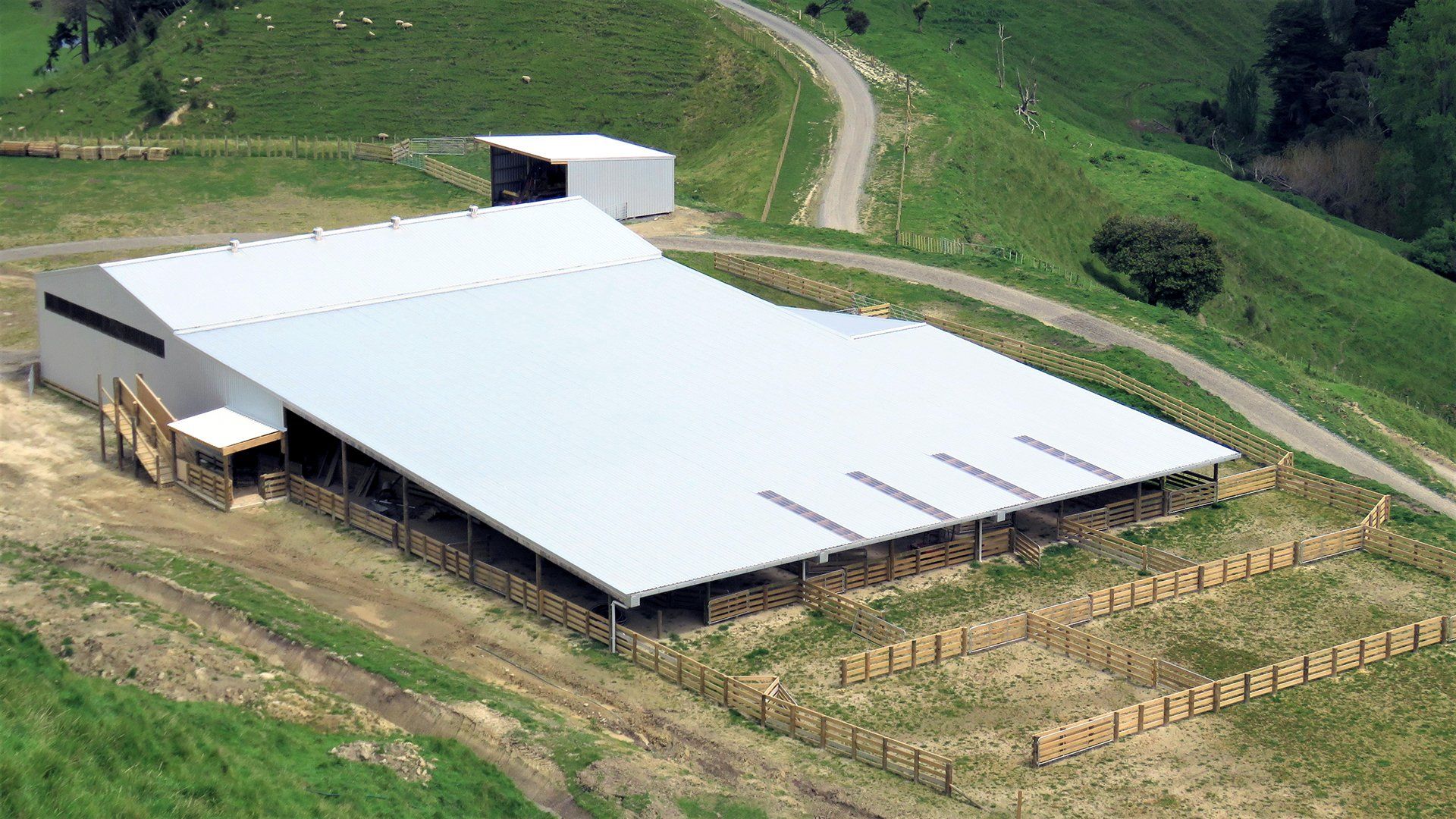 An aerial view of a large white-roofed barn on a grassy hillside, with hay bales stacked in a rectangular pattern nearby.