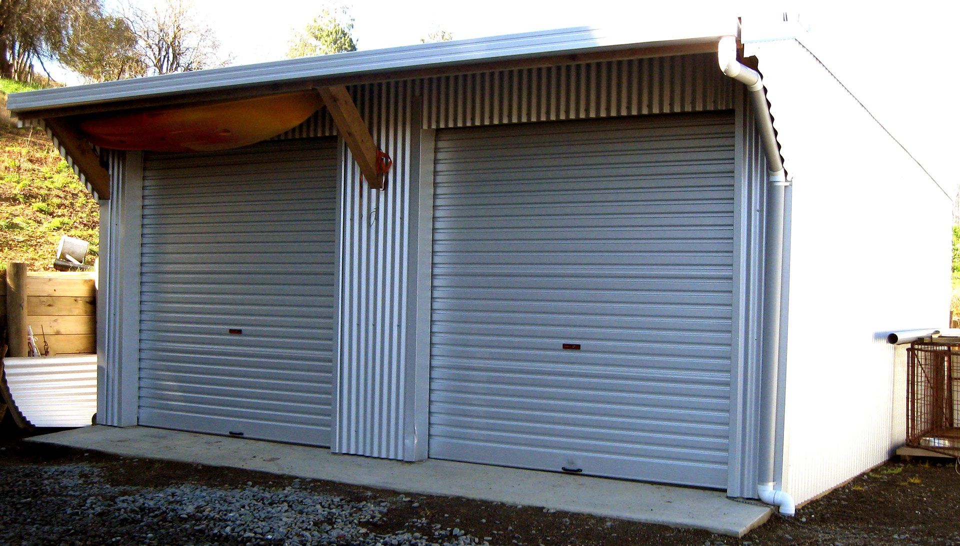 A double garage with two gray roller doors, a corrugated metal facade, and a concrete driveway in a rural setting.