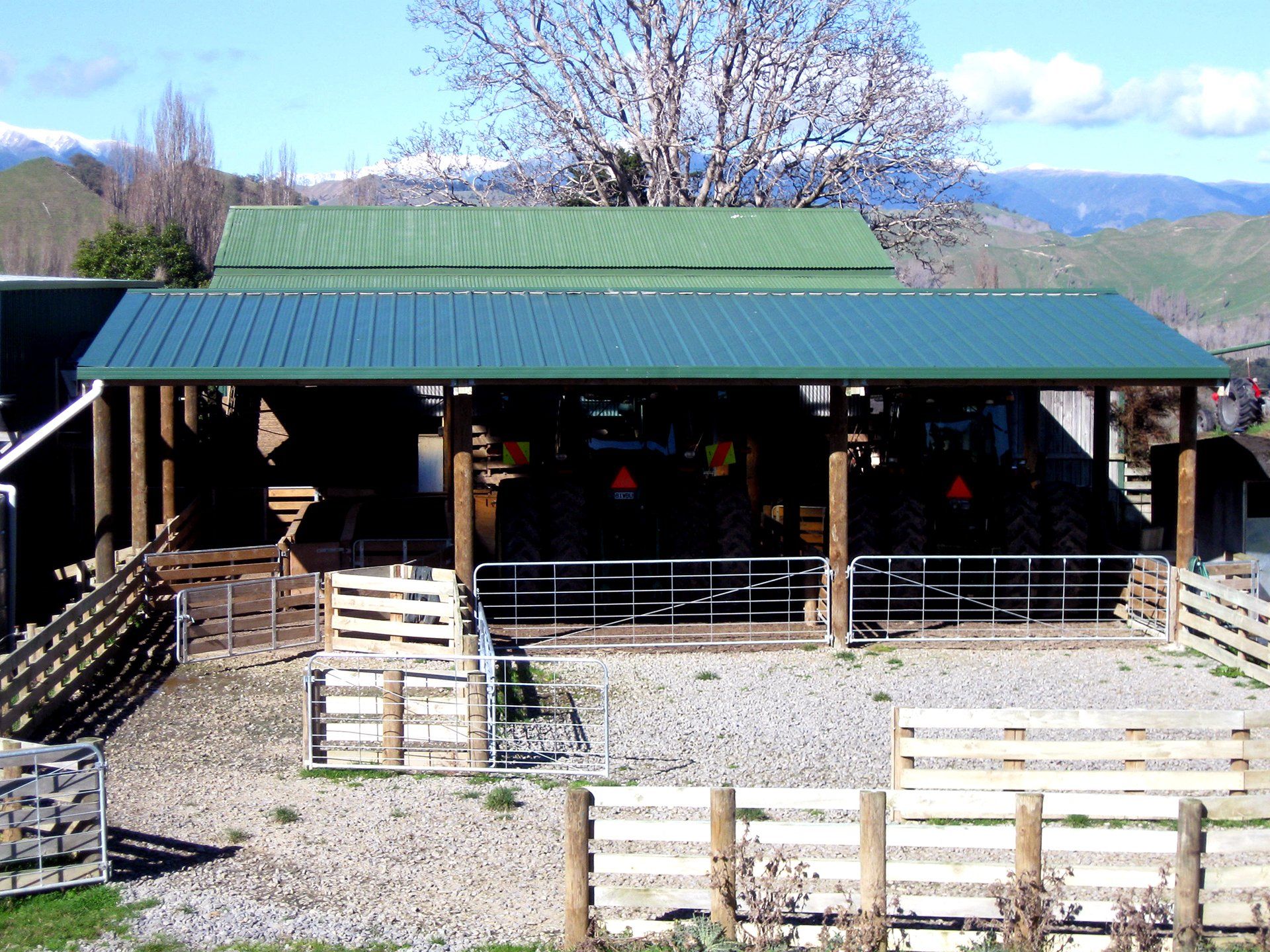 A farm building with a green metal roof, open front, and fenced gravel paddock with mountains in the background.