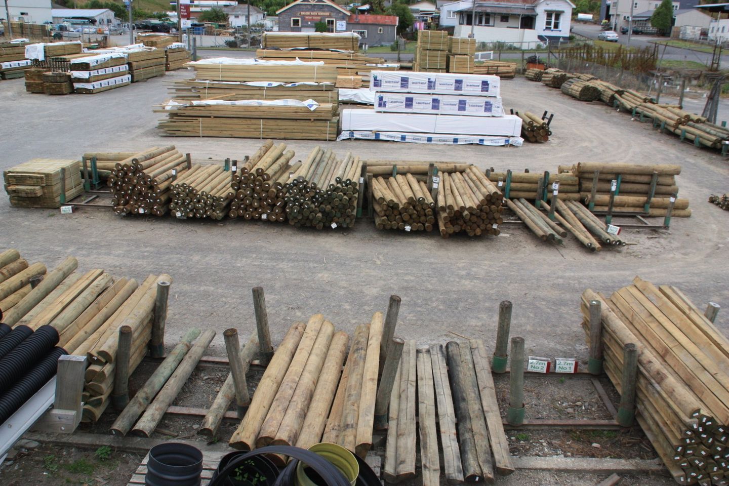 An outdoor lumber yard displaying various piles of wooden fence posts, planks, and timber materials on a gravel lot.