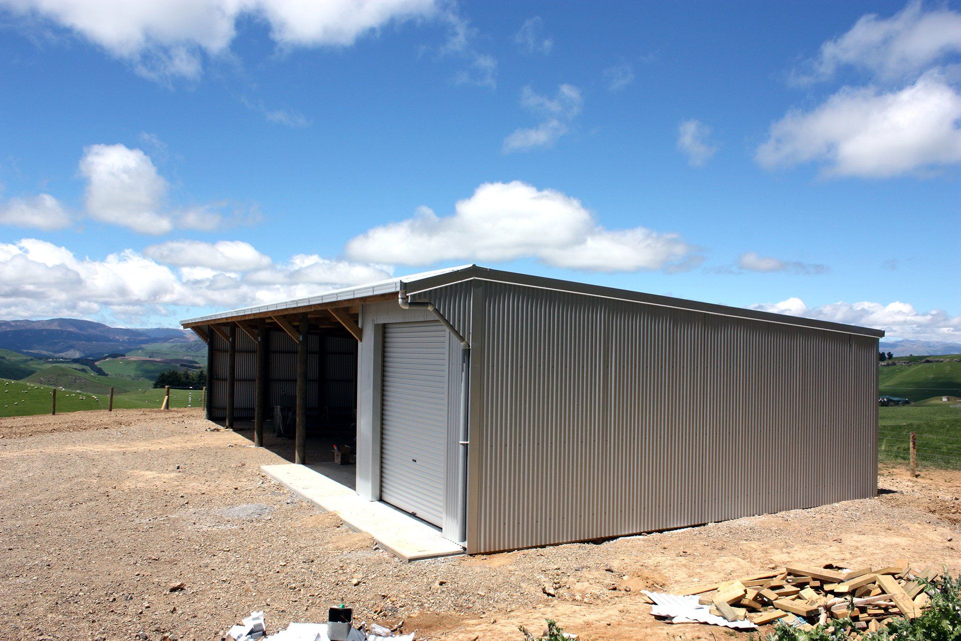 A beige, corrugated metal garage with an open side bay stands on a gravel lot under a bright, cloudy blue sky.