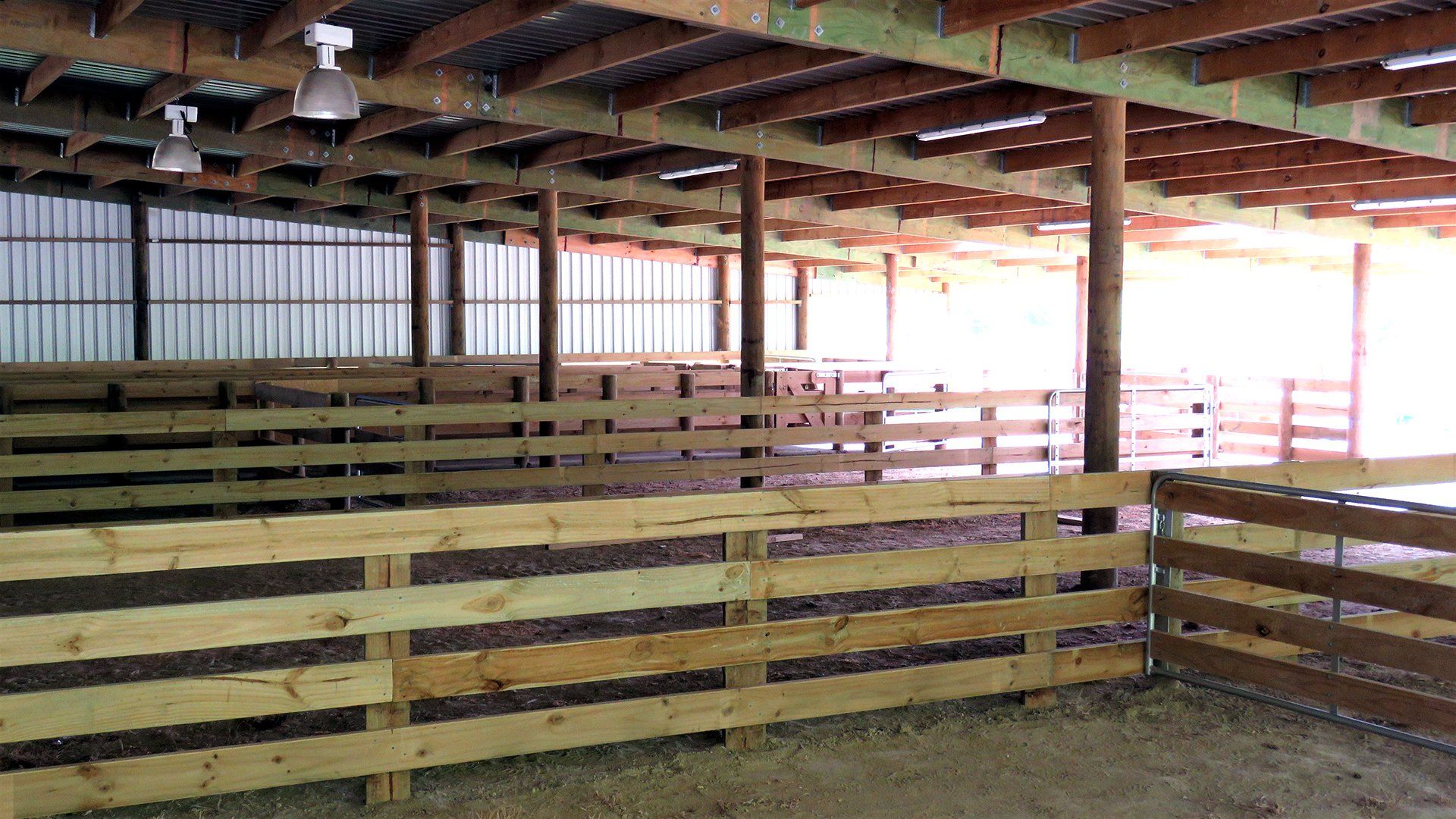 An indoor livestock barn with wooden rail fences creating several stalls under a metal roof.