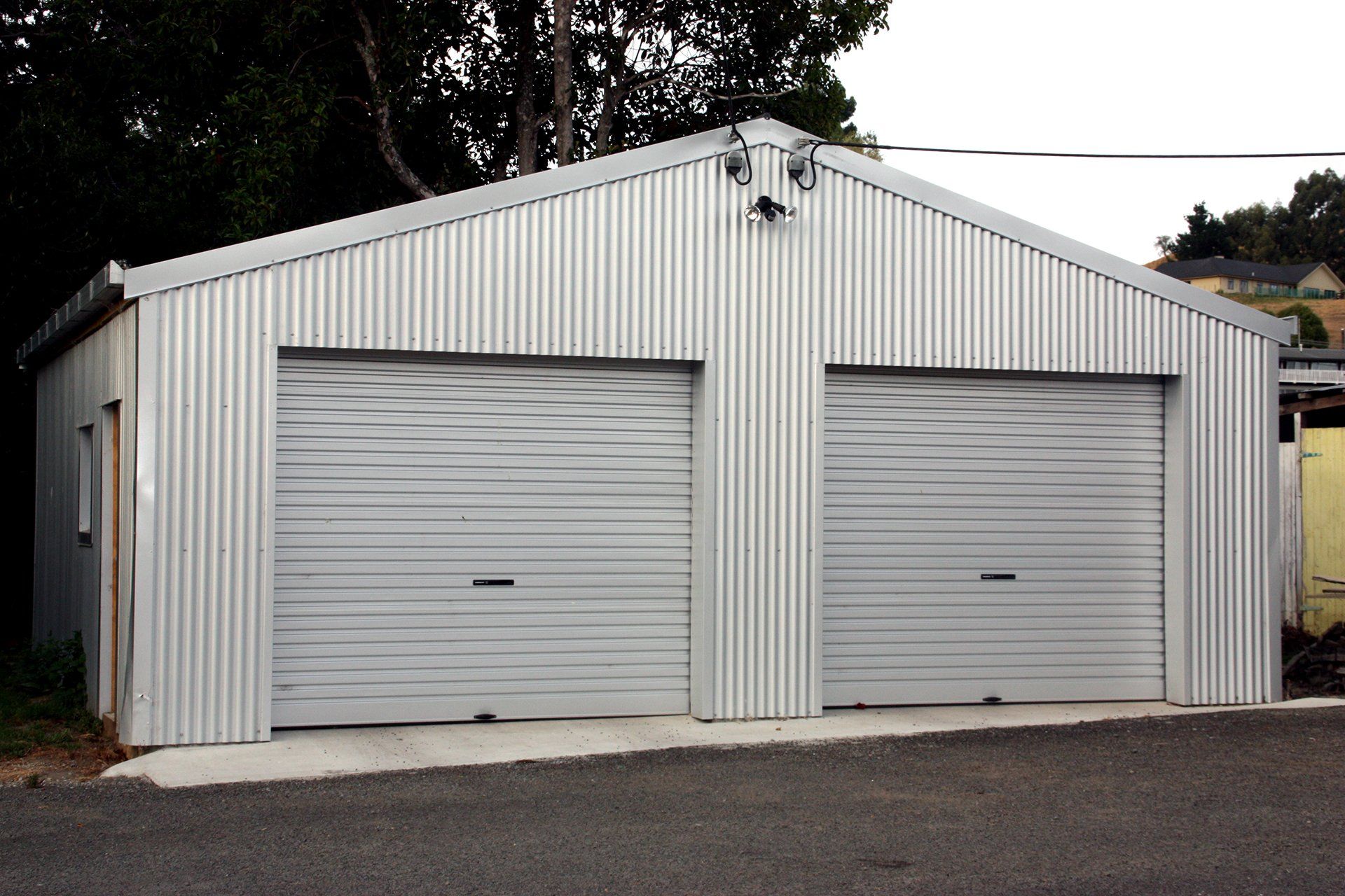 A detached, silver corrugated metal double garage with two roller doors, situated outdoors on a gravel lot.