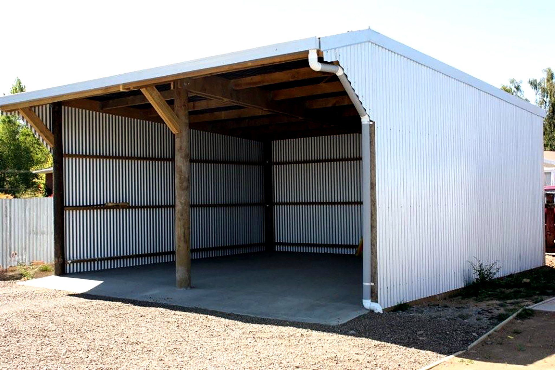 A silver, metal-sided carport with a concrete floor and wooden support posts on a gravel lot.