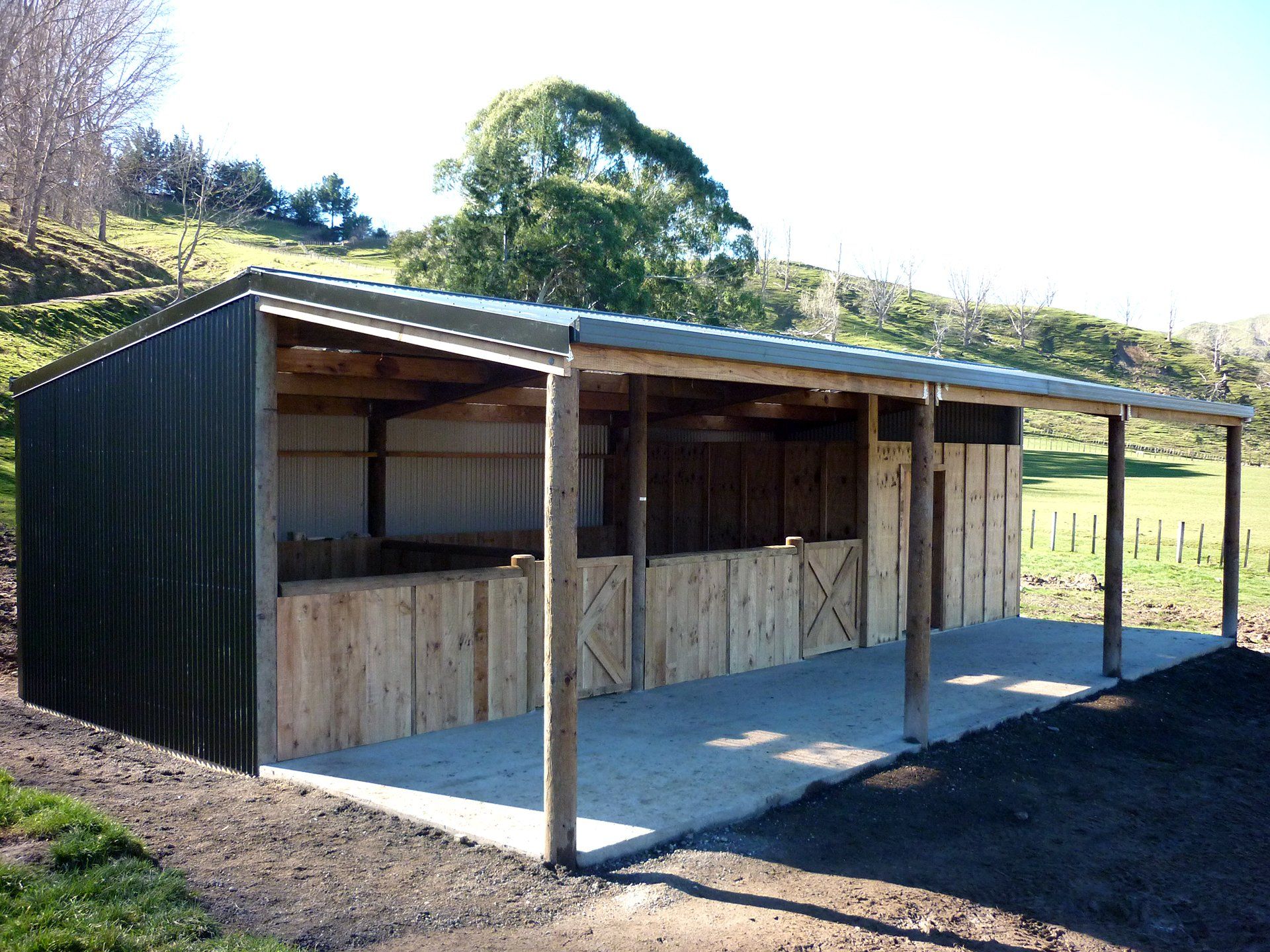 A three-sided, green metal-roofed animal shelter with wooden stalls, situated on a concrete pad in a grassy field.