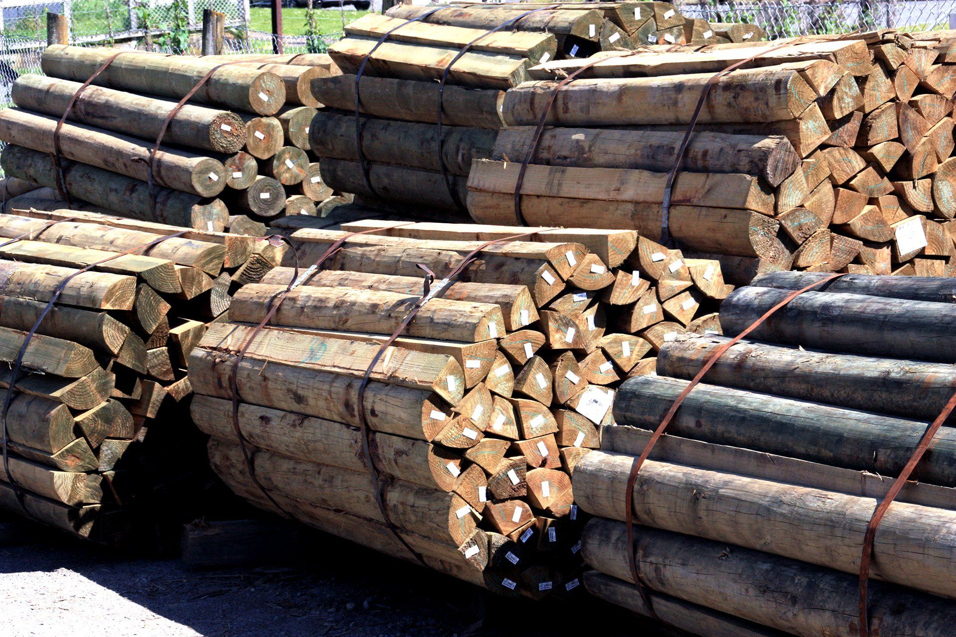 Bundles of stacked, cylindrical wooden posts secured with metal strapping in an outdoor storage yard.