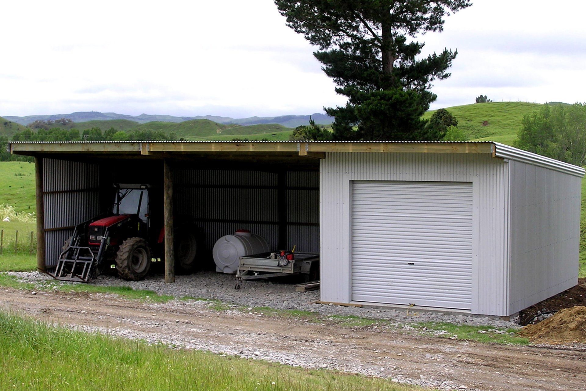 A rural pole shed with an open bay housing a red tractor and a closed bay with a white roller door on a grassy lot.