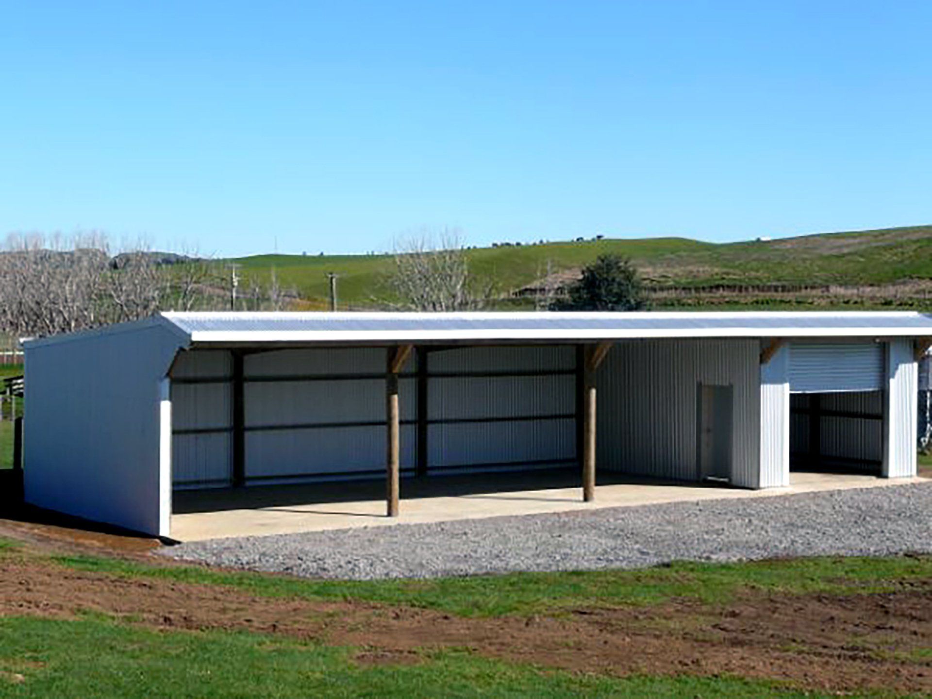 A white pole barn with an open-sided carport section and a closed garage bay, set on a gravel pad in a grassy landscape.