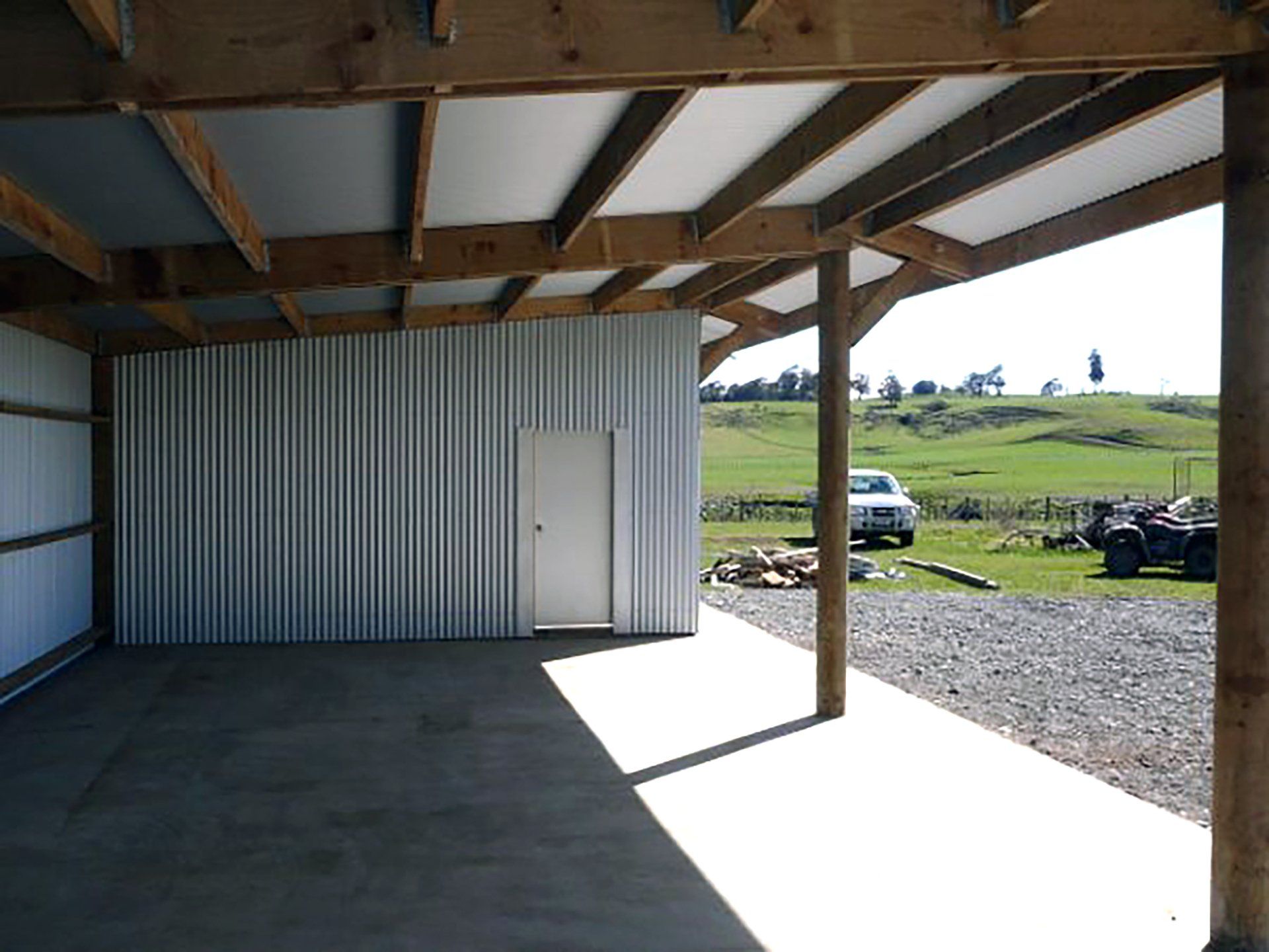 A shaded, concrete-floored structure with a white corrugated wall and a door, overlooking a rural landscape with a vehicle.